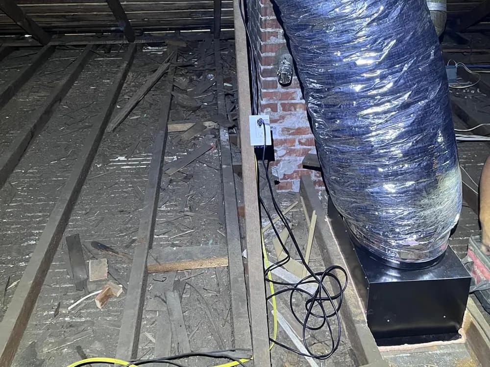 Carcasa de ventilador de ático montada sobre la estructura y conectada a un tramo de ducto flexible aislado.