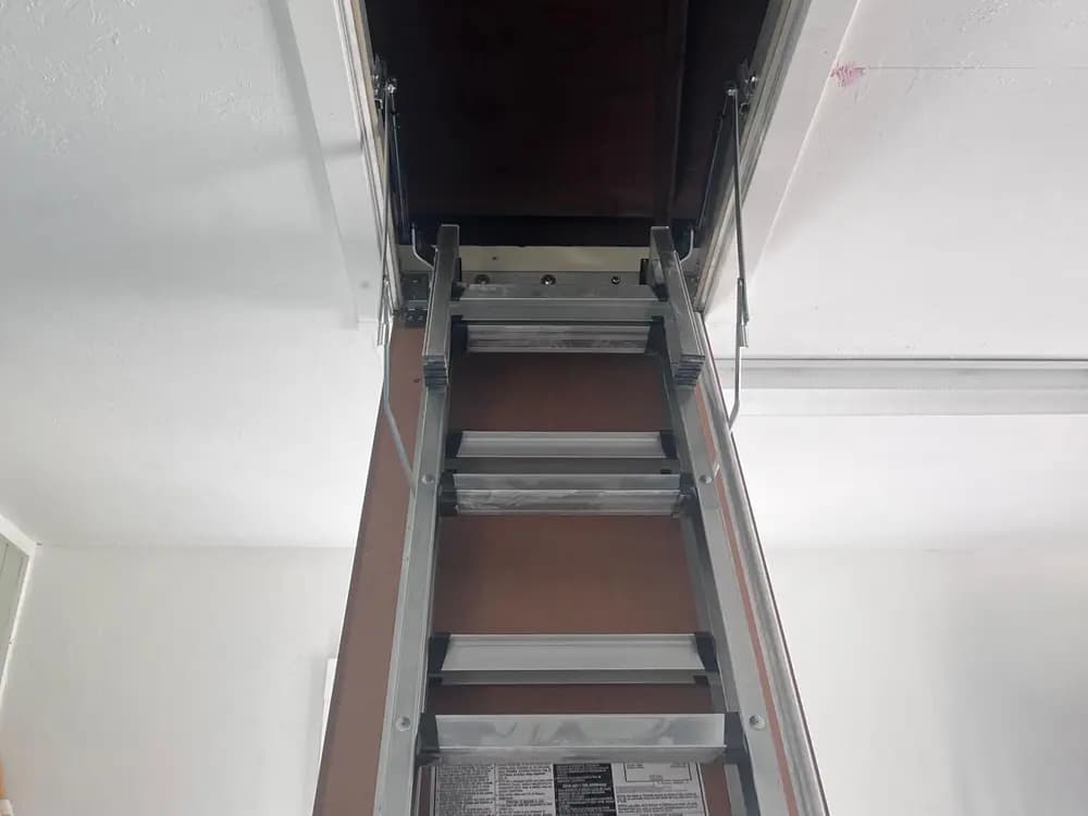 Attic ladder door hinged open against its framed ceiling opening, hinge arms visible at each side and the aluminium ladder sections extending down into the room.