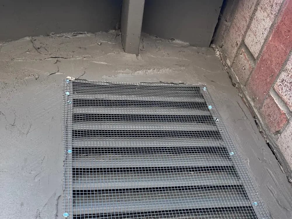 Rejilla de cimentación cubierta con malla galvanizada, fijada en los bordes y adherida con mortero al muro circundante.